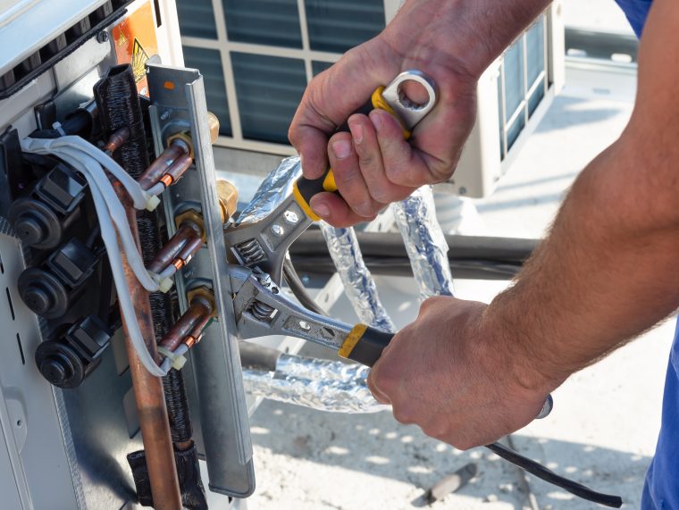 Un technicien CVC travaille sur des unités de climatisation sur le toit d'un nouveau bâtiment industriel. Le technicien utilise une clé fixe pour serrer l'unité extérieure de climatisation. L'homme tient une clé à la main.